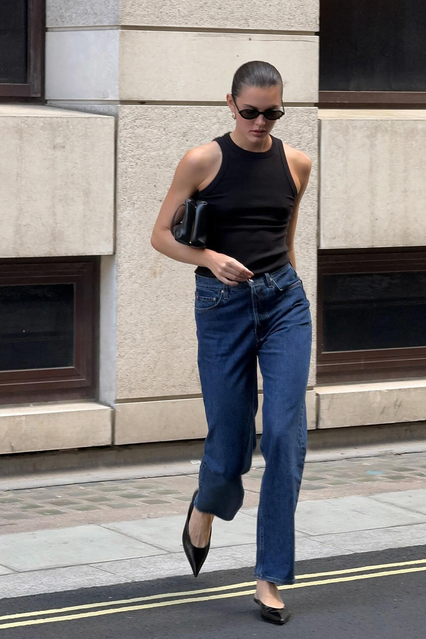Denim and shoe combos shown in a street style photo of a woman crossing the street wearing a black halter neck tank top with puddle jeans, black pointed mules, black sunglasses, a black handbag, and gold earrings