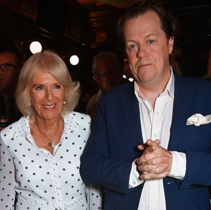Queen Camilla wearing a white polka-dot dress standing next to her son Tom Parker Bowles and smiling
