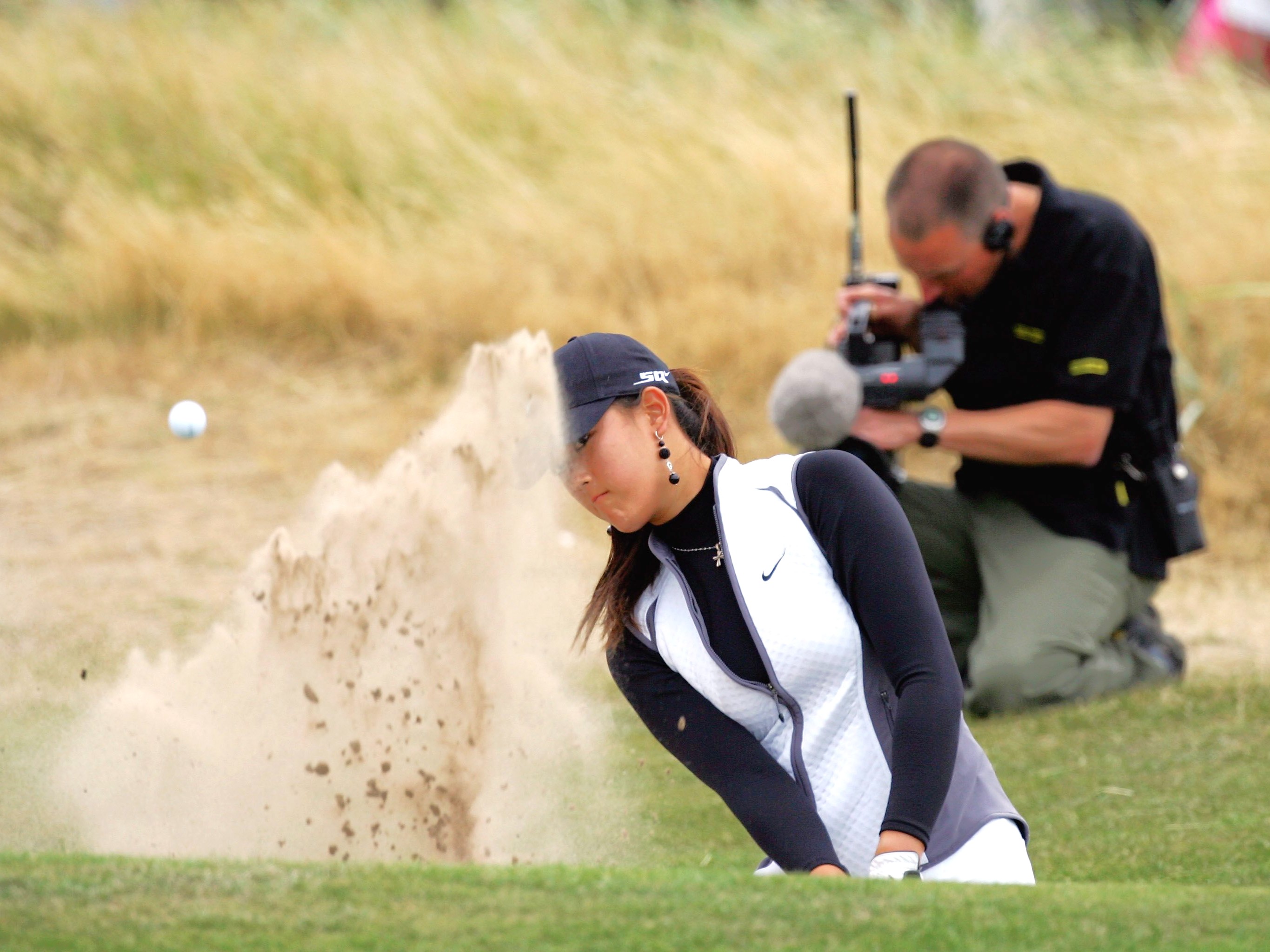 Michelle Wie playing from a bunker with a cameraman kneeling behind the bunker filimng the shot with a hand-held camera