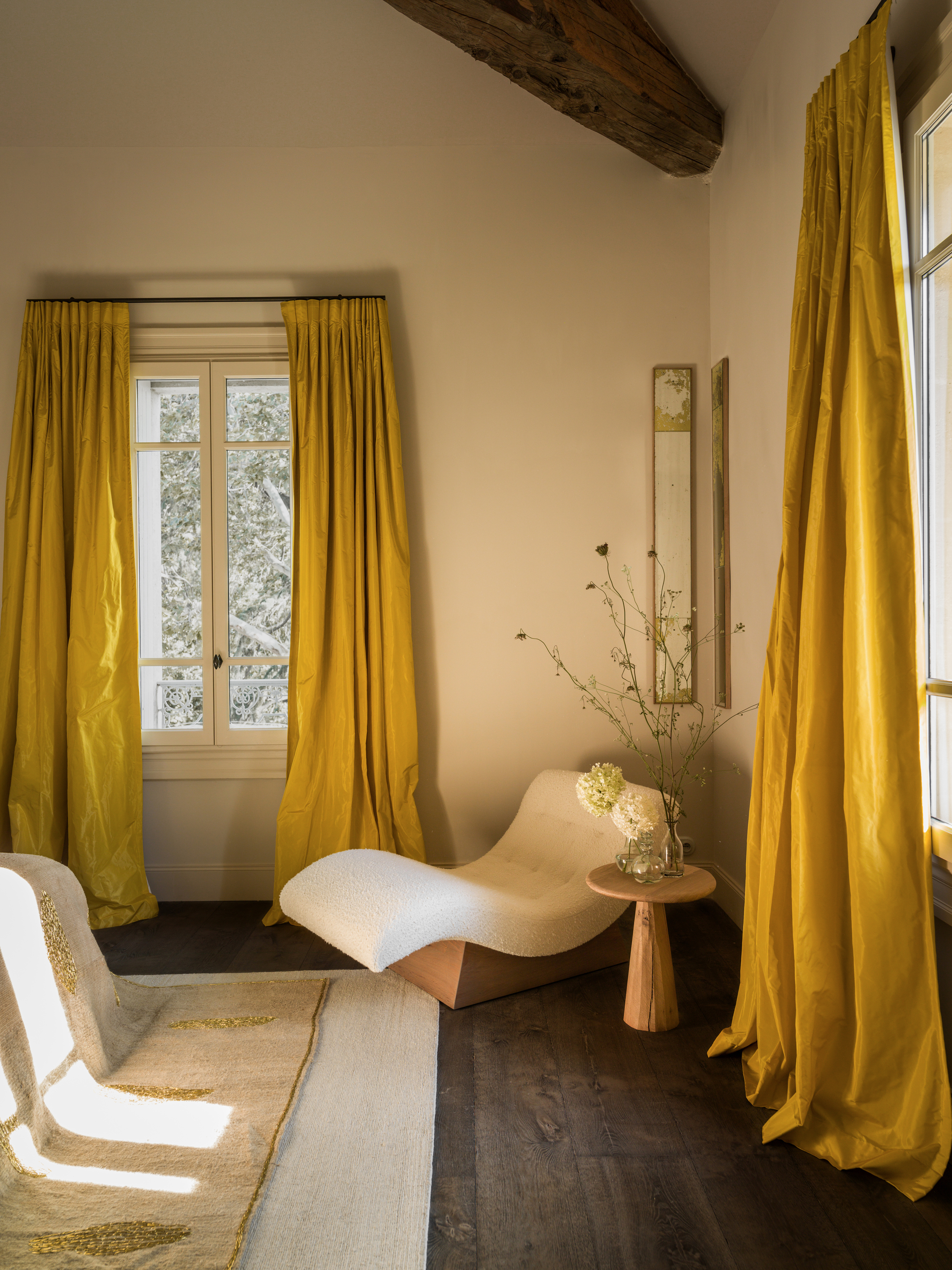 A white bedroom with dark wooden floors and two windows that have heavy, sunflower yellow curtains on them. There is a modern, white boucle accent chair with a small wooden side table next to it.