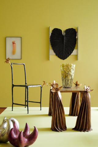 In a yellow-painted showroom, playfully designed furniture including a chair and a coffee table appear alongside wall hart and creative sculptural pieces.