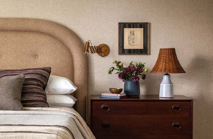 A bedside table styled with lamp, painting, wall light and flowers