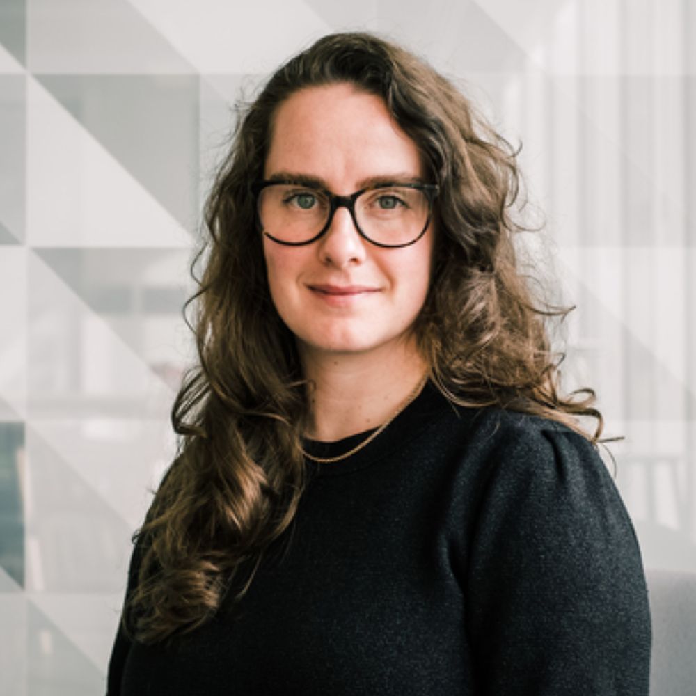 A woman with long, wavy brown hair and black glasses looks confidently toward the camera. She wears a simple black top and a gold necklace.
