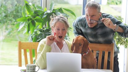 An older couple look shocked as they look at their laptop screen at the dining room table. 