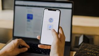 A person holds their iPhone and unlocks their 1Password account using Face ID. In the background is a laptop.