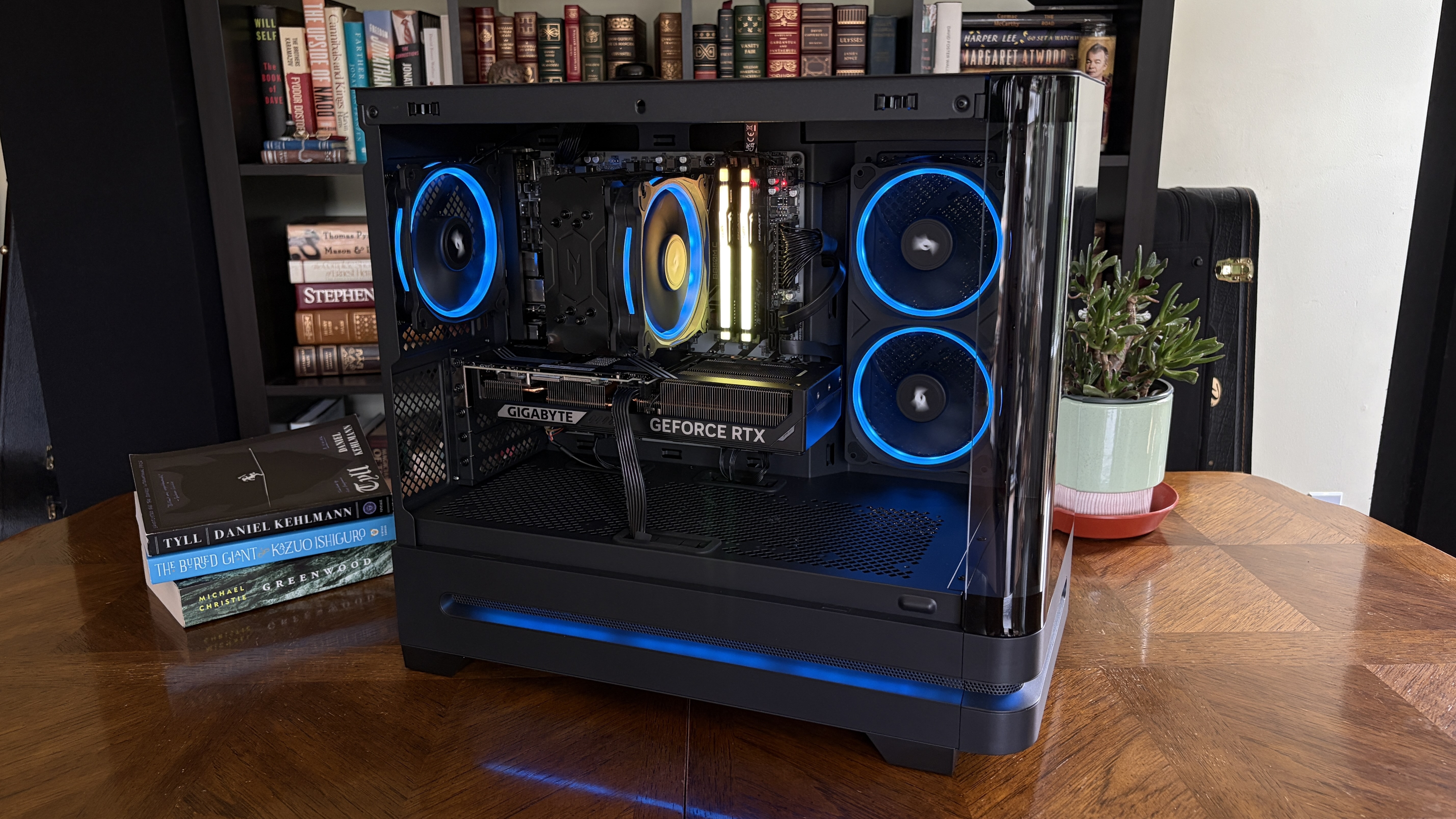 A modern Nitro 65 computer tower with glass panels, illuminated blue fans, and a GIGABYTE GeForce RTX graphics card, placed on a wooden table with several books and a green potted plant.