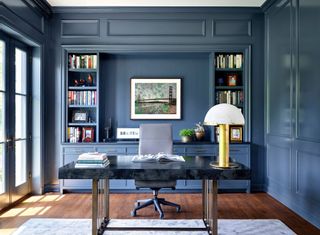 home office with blue walls and large black topped desk and contemporary table lamp and desk chair with painting and shelves behind