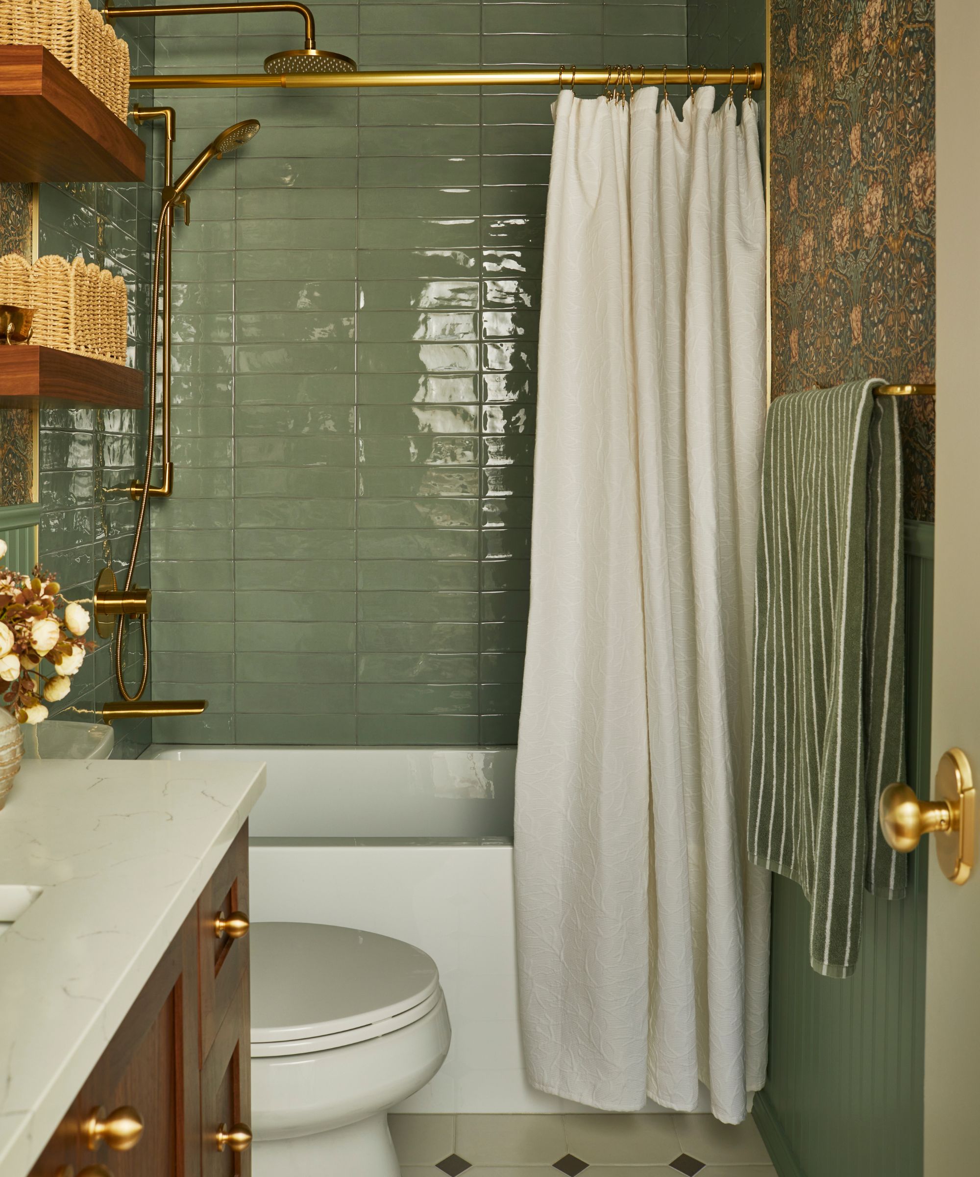 a sage green tiled bathroom with pink and green floral wallpaper, a wooden vanity, bath, and floating shelves