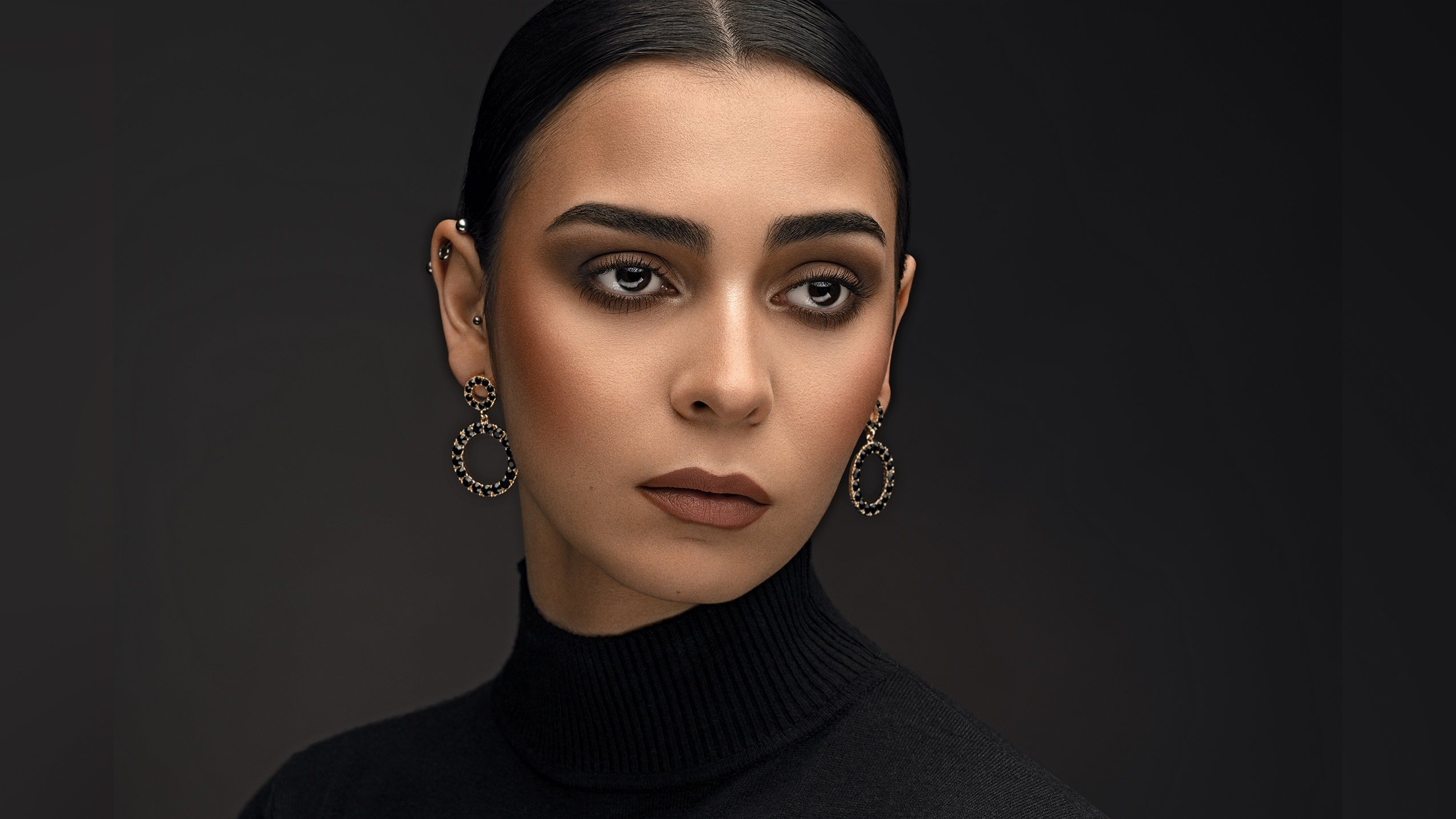 A woman in a black turtleneck and large circular earrings poses against a dark background, exuding elegance and confidence