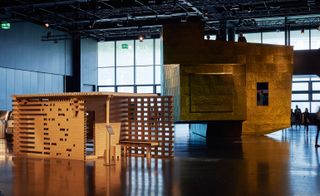 View of the 'Design at Large' exhibition featuring Shigeru Ban's Paper Tea House - a structure made out of paper and cardboard with a flat roof and waiting area. There is also a gold, irregular shaped structure with a rooftop terrace beside it. Both structures sit in a space with dark walls, windows and black floors