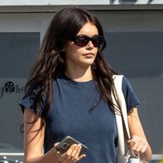 Kaia Gerber wearing sunglasses with a blue T-shirt and black skirt-over-pants look.