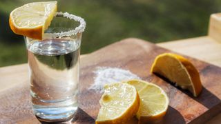 Shot of tequila with salt around the rim and lemons on a wooden chopping board