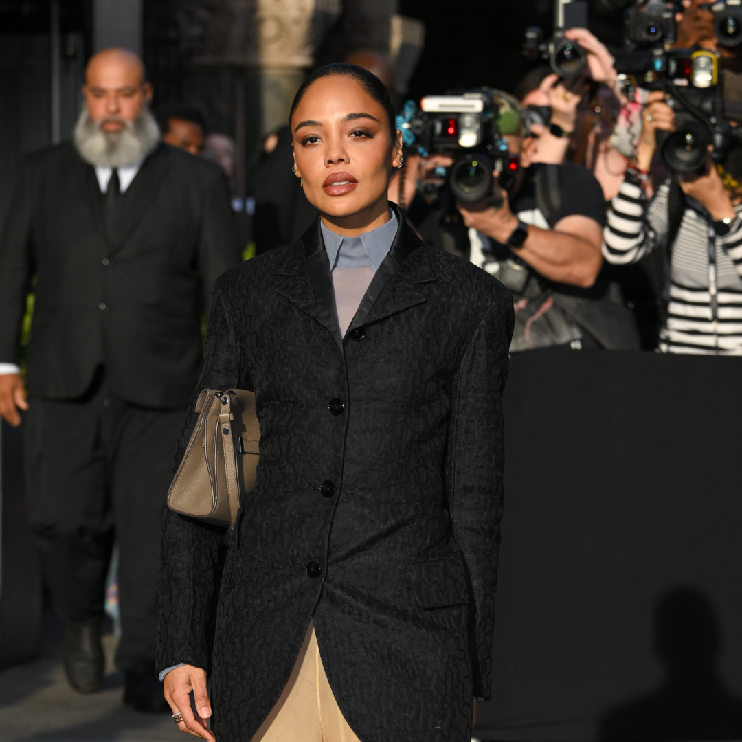 A woman with a sleek bun hairstyle wearing a blazer and collared shirt while holding a taupe colored bag. 