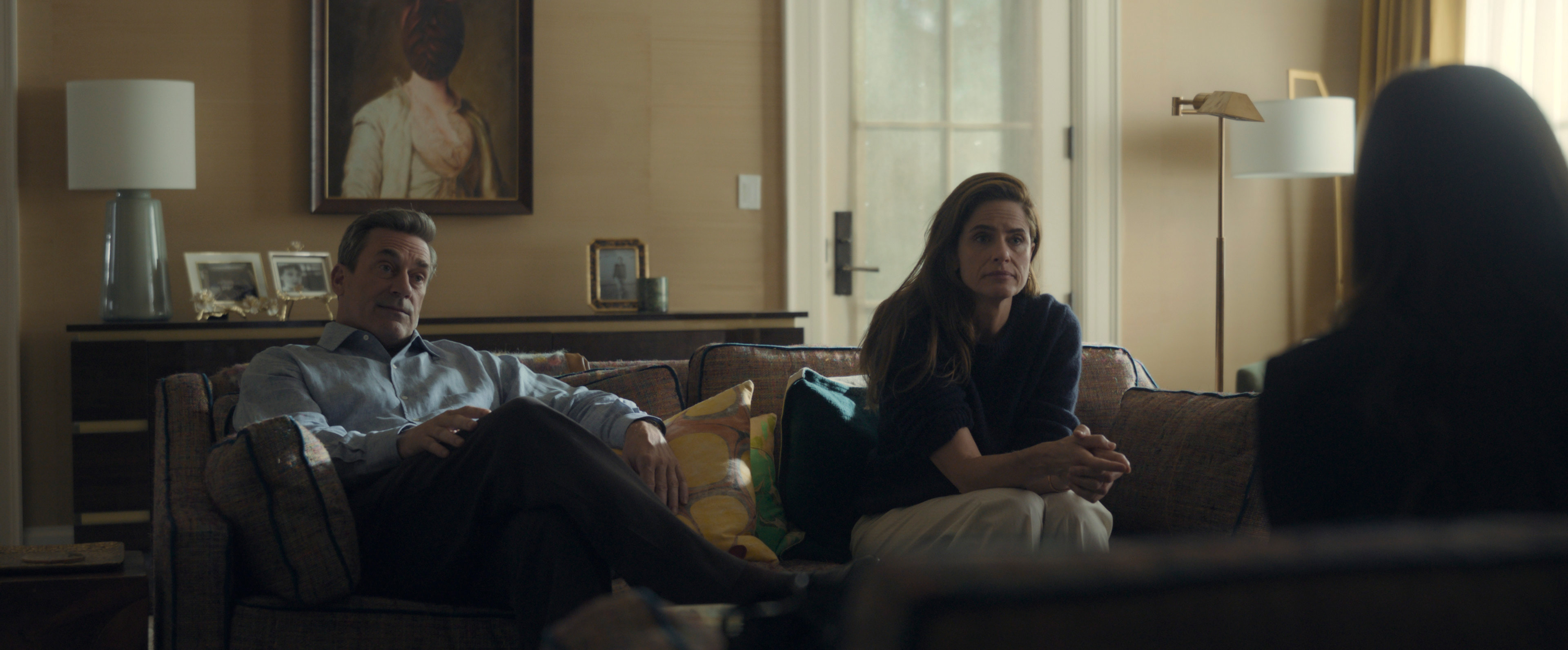 amanda peet as mel cooper and jon hamm as andrew cooper sitting on a couch having a tense conversation in your friends and neighbors season 2