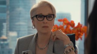 A shot of Miranda Priestly (Meryl Streep) sitting at a desk in New York with a view of the city behind her. Flowers can be seen in the windowsill. 