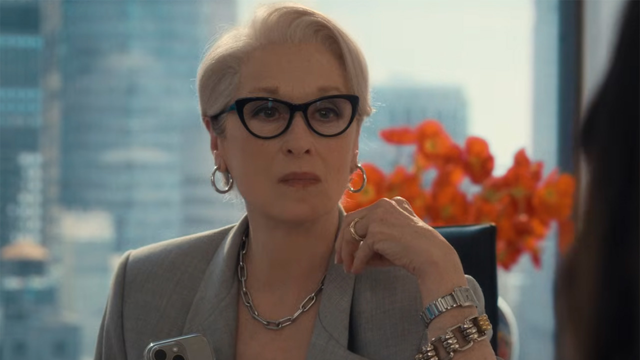 A shot of Miranda Priestly (Meryl Streep) sitting at a desk in New York with a view of the city behind her. Flowers can be seen in the windowsill. 