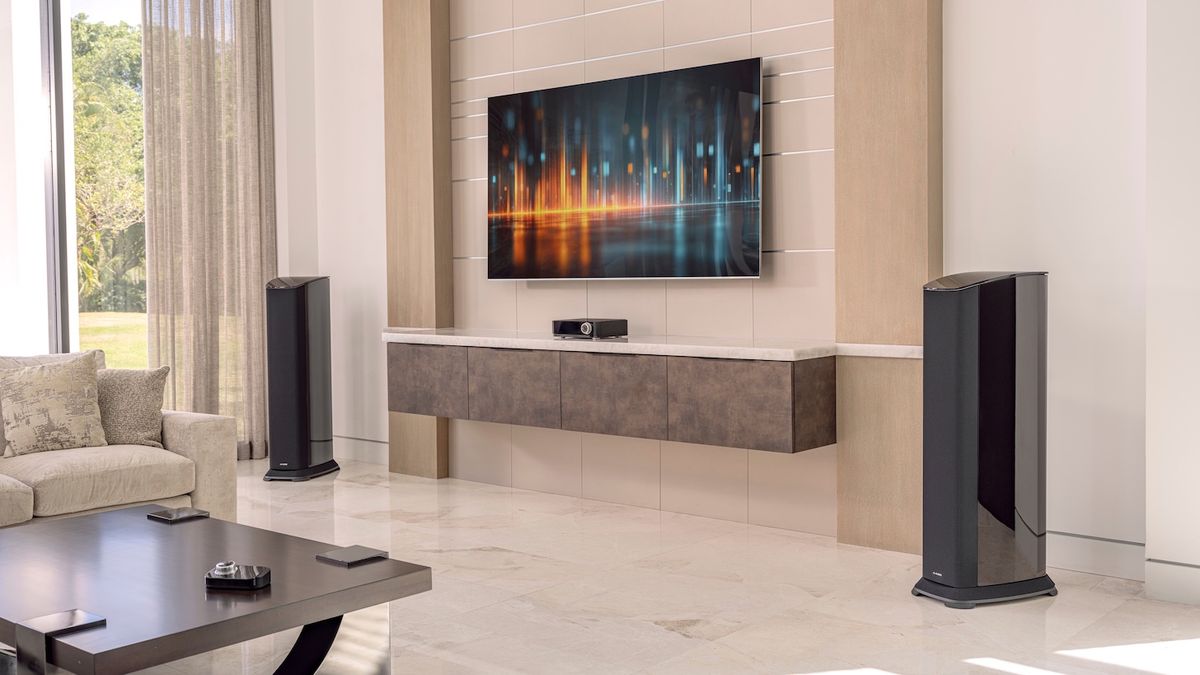 Two JL Audio Primacy floorstanding speakers, a Centrepiece unit and a wireless controller in a modern lounge setting with a wall-mounted TV.