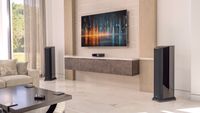 Two JL Audio Primacy floorstanding speakers, a Centrepiece unit and a wireless controller in a modern lounge setting with a wall-mounted TV.