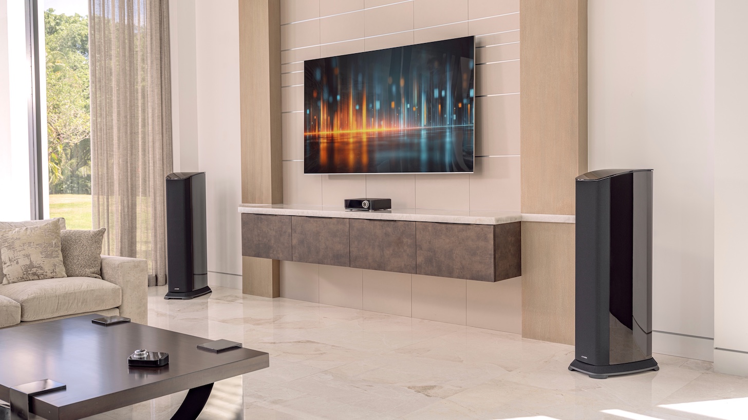 Two JL Audio Primacy floorstanding speakers, a Centrepiece unit and a wireless controller in a modern lounge setting with a wall-mounted TV.