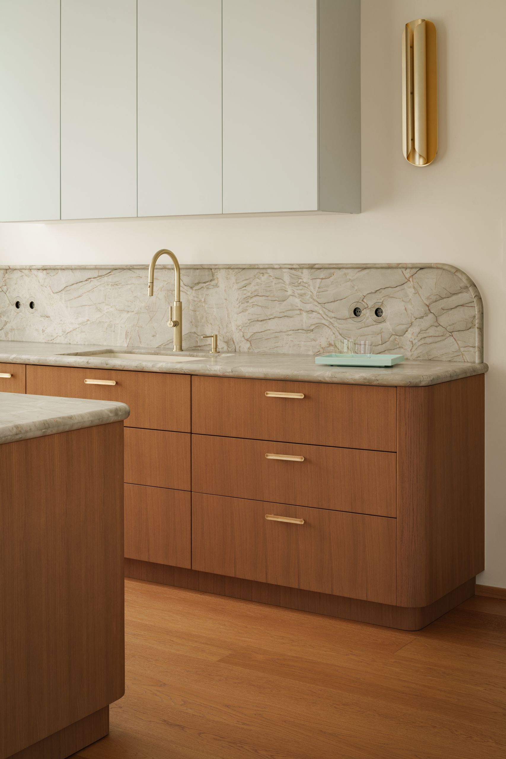 Image of a kitchen with warm wooden cabinetry that has green and gray marble countertops, light blue floating cabinets, and gold hardware and lighting.