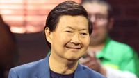 Ken wearing a blue suit and smiling at 99 To Beat competitors in Season 2. A man behind him is wearing a green shirt. 