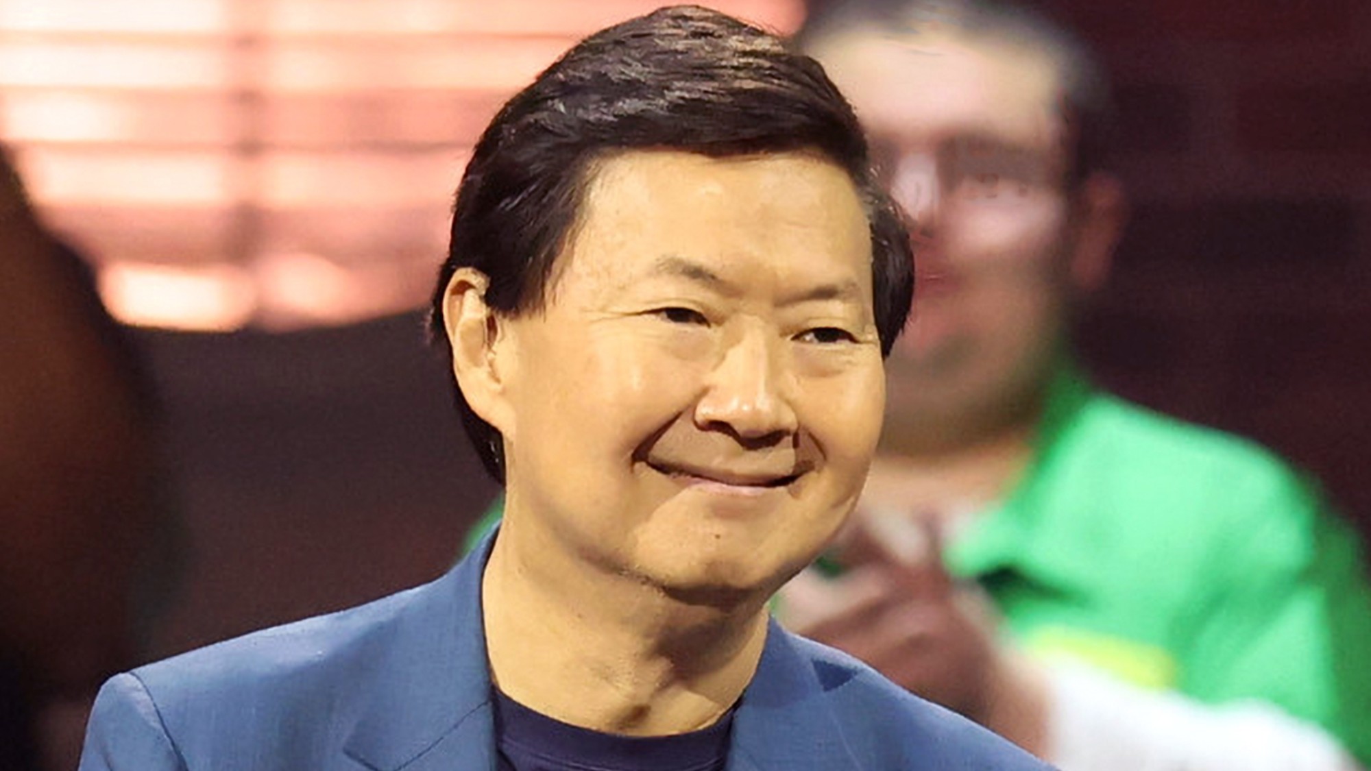 Ken wearing a blue suit and smiling at 99 To Beat competitors in Season 2. A man behind him is wearing a green shirt. 