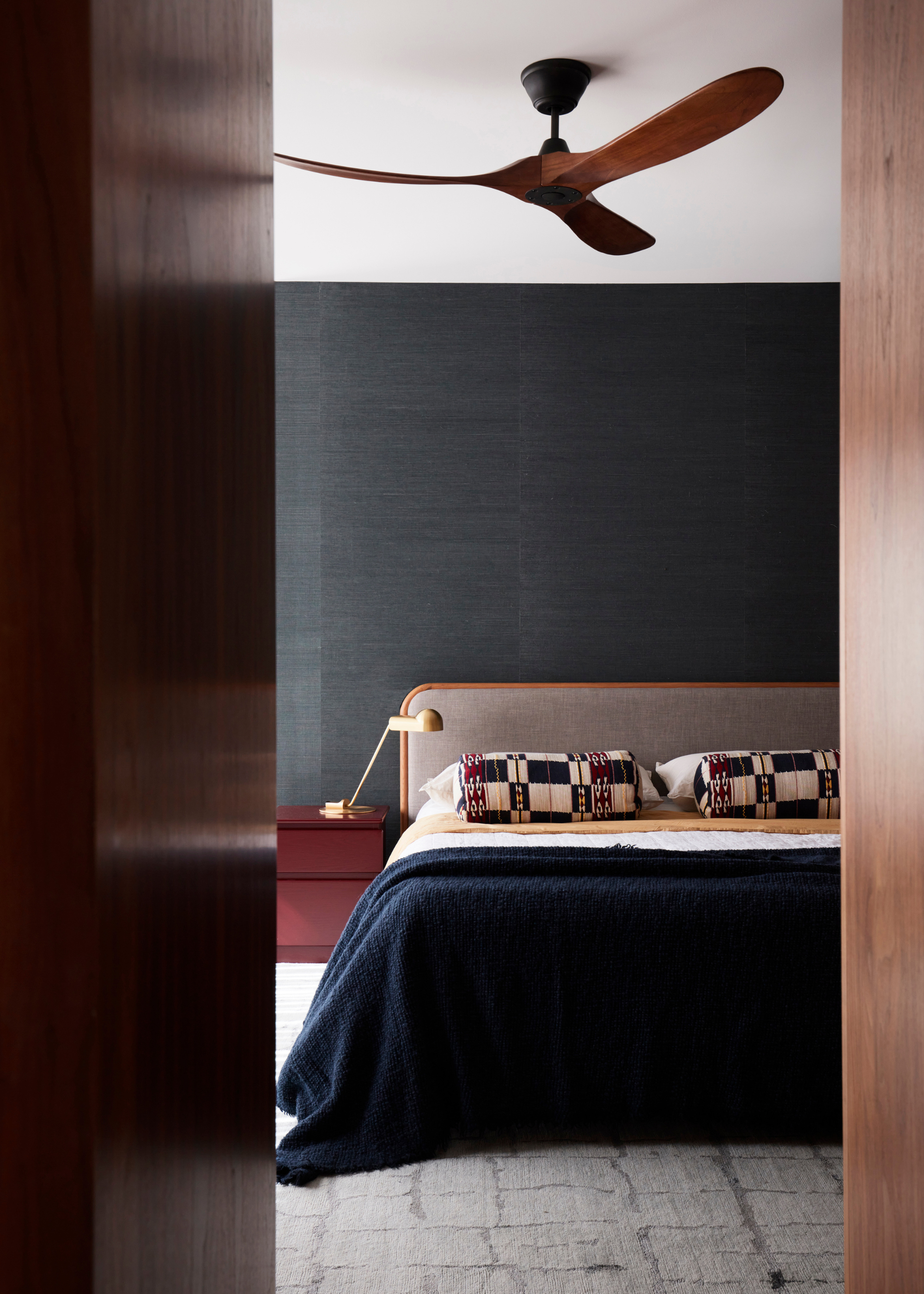 A cool-toned bedroom with bolster pillows, a maroon side table, a wooden ceiling fan and a headboard