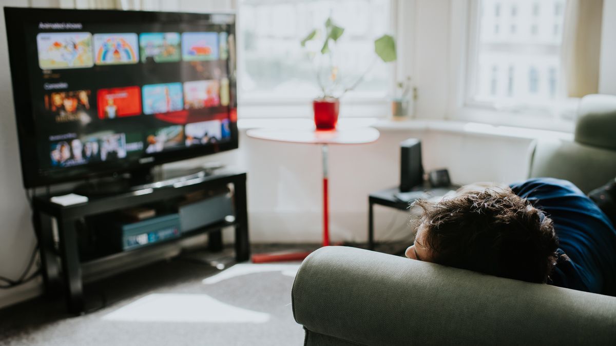 A man lies on a sofa, staring at a large, flatscreen TV displaying a choice of shows on a streaming platform