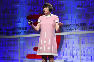 Alice is wearing a pink midi tunic dress which has a white border at the arms, neck and bottom, and white rectangles and circles embroidered from the bottom to the top. Alice is using her arms whilst speaking on stage.
