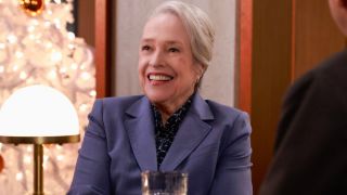 Kathy Bates sits smiling at a table with a drink in front of her in Matlock S1 E7 - 