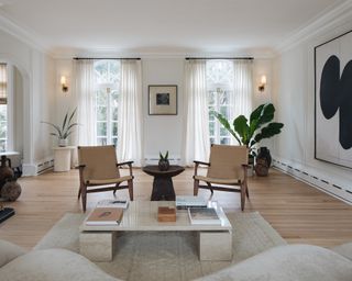 Neutral living room with coffee table, armchairs, area rug, large artwork and two windows dressed with full length curtains