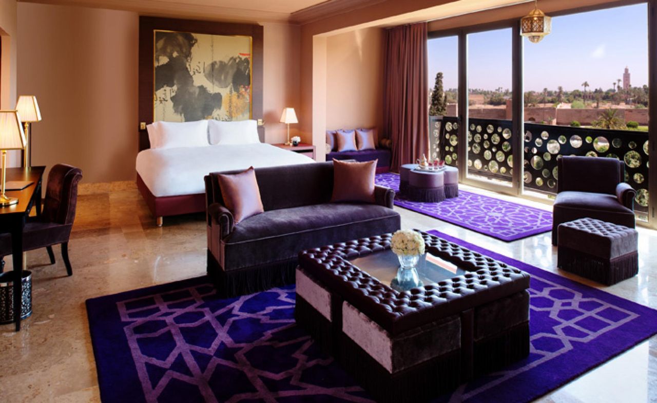 A hotel bedroom with a bed, a wooden side table, a lamp, a purple sofa, a wooden glass top coffee table, a desk, a purple rug and large sliding doors.