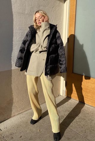 a roundup of the best puffer jackets shown in a photo of a woman wearing a black puffer coat over a tan blazer with yellow trousers and black boots