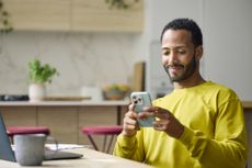 A smiling young man in a yellow sweater holds his phone and smiles, with a laptop and cup of coffee in front of him.