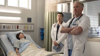 L-R: (Joel Kim Booster) and Dr. Cox (John C. McGinley) standing next to a bedbound patient in the hospital in an episode of "Scrubs" season 10