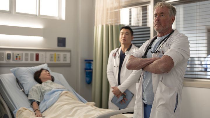 L-R: (Joel Kim Booster) and Dr. Cox (John C. McGinley) standing next to a bedbound patient in the hospital in an episode of "Scrubs" season 10