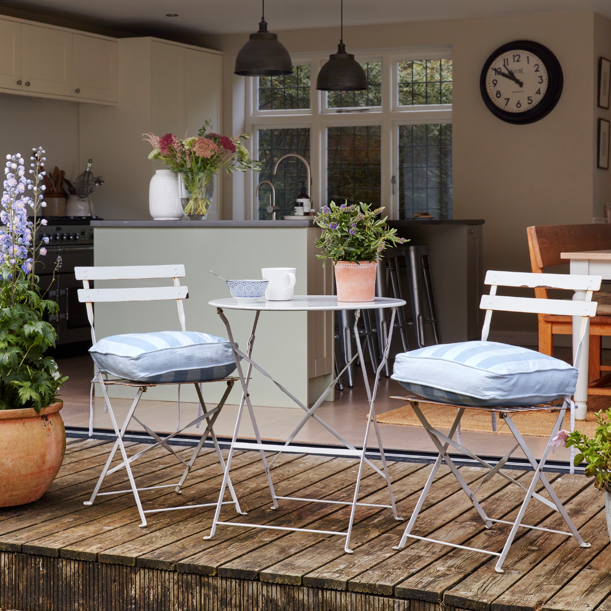 A patio with a white bistro set with striped seat cushions and potted plants on the table and around the set