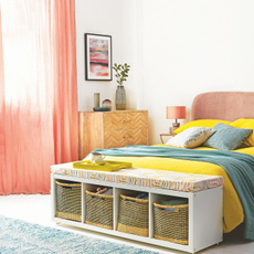 Bedroom with white walls, double bed with yellow and green bedding, stool with storage baskets at the end of the bed.