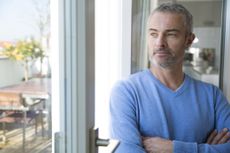 An older, attractive man looks out the window or french door to a patio. His arms are folded and he appears thoughtful.