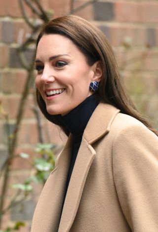 Kate wears a tan coat, blue earrings, and a smile at an engagement