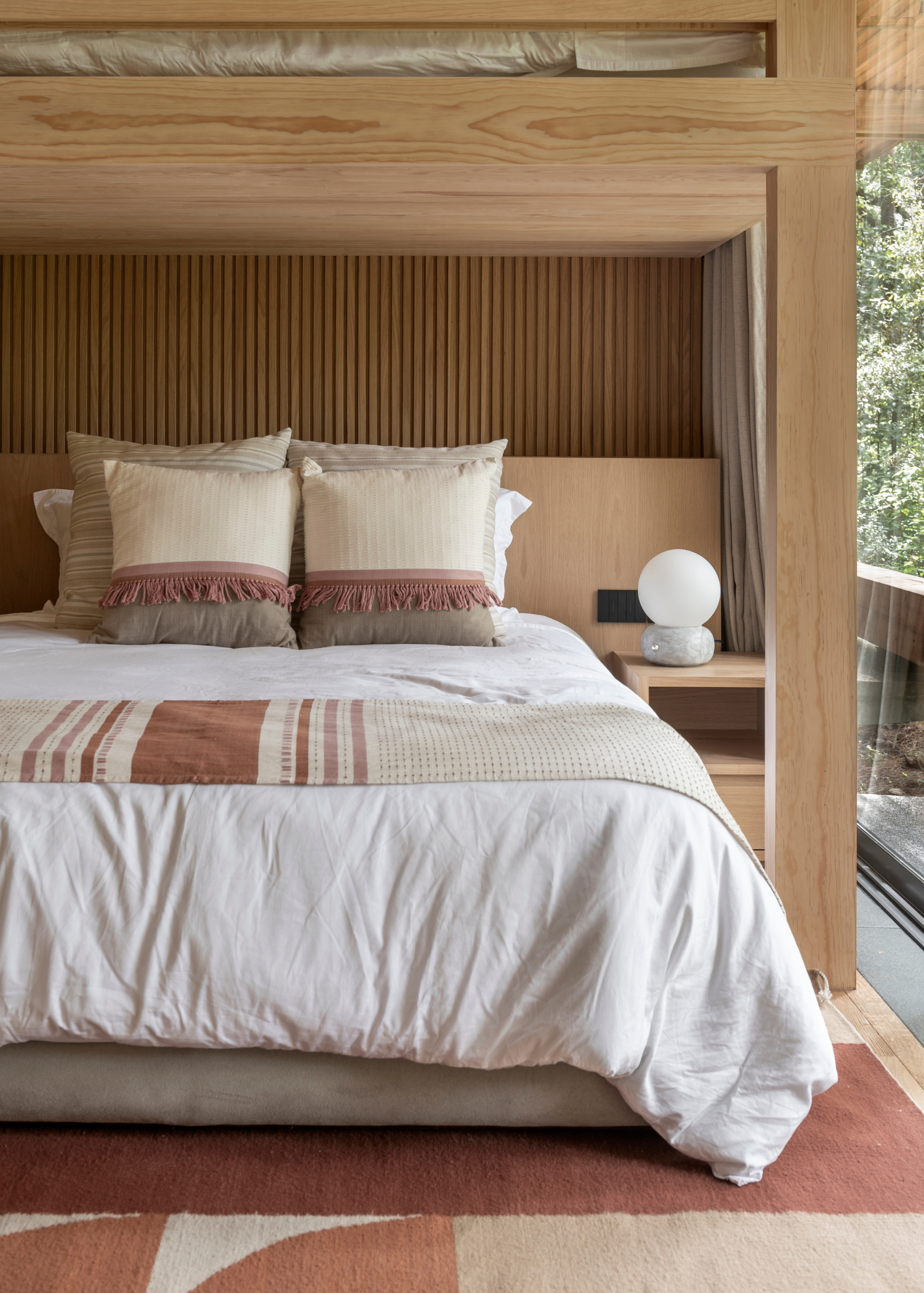 A warm bedroom with wood paneling, a side table, and a white bedscape with textured colorful pillows