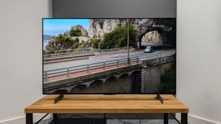 The LG C4 42-inch OLED TV straight-on on a TV stand. On screen is a car driving out of a tunnel in mountainous terrain.