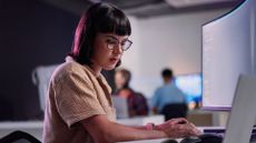 Female software developer working at a desktop computer in an open plan office space with laptop next to screen.
