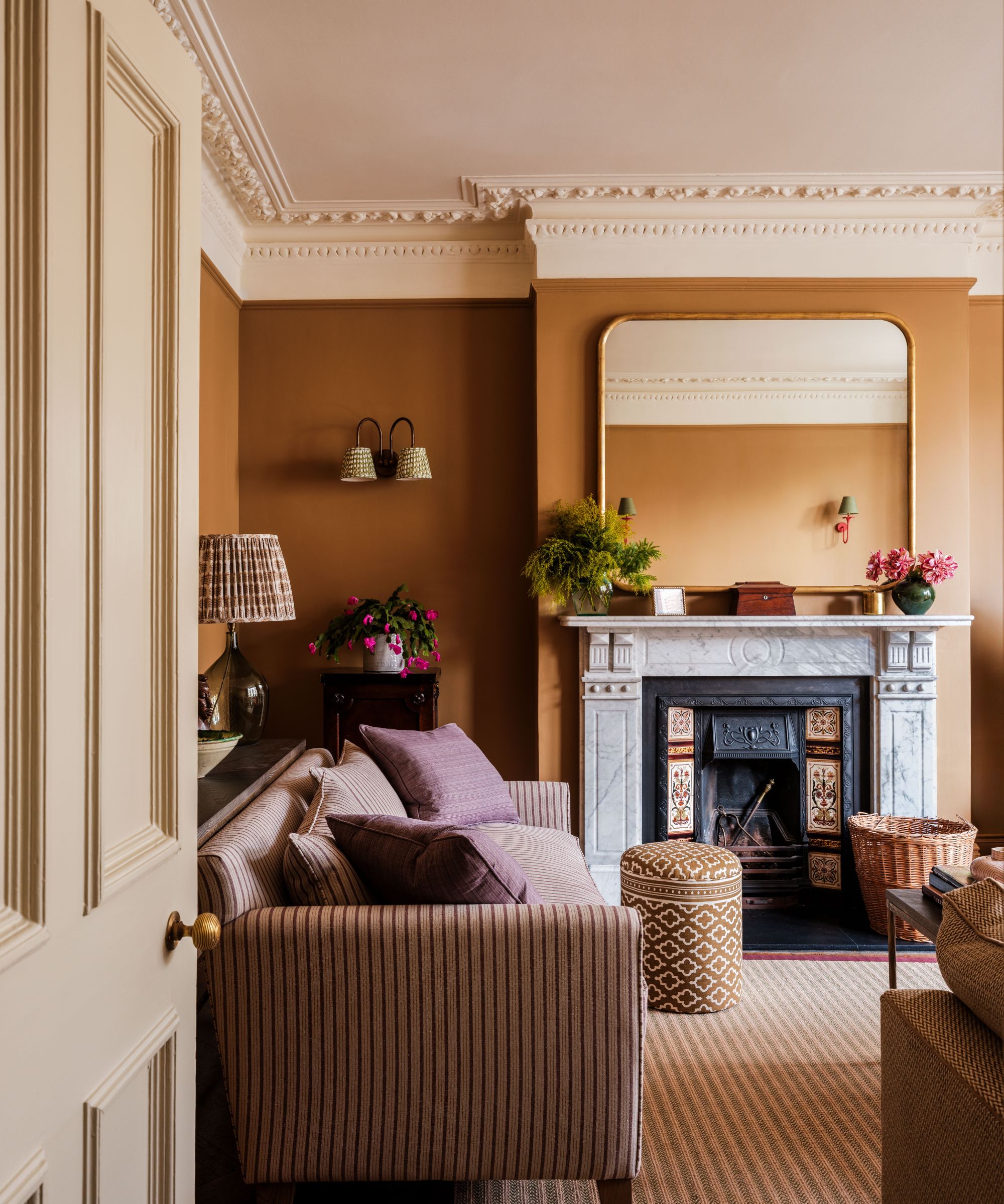 Living room with brown walls, a marble Victorian fireplace, a large gold mirror above, a striped purple sofa, a striped jute rug, and a patterned ottoman.