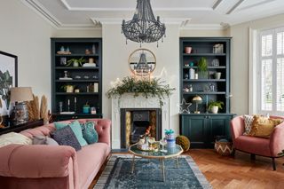 Edwardian living room with neutral walls, dark green painted open shelving in alcove, pink sofa and armchair, chandelier, and Christmas garland over fireplace