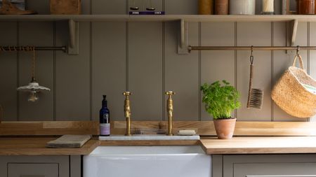 A farmers sink with gold faucets, set into a wooden countertop. Greige wall panelling behind, leading to matching shelving. 