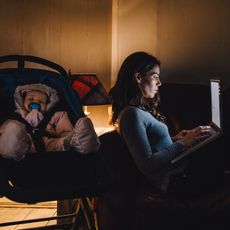 A mother working in the dark on her laptop while her baby sleeps in a stroller.