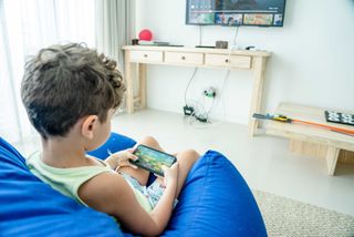 A kid playing a mobile game on a smartphone, with a TV displaying a gaming interface on the wall in the background.