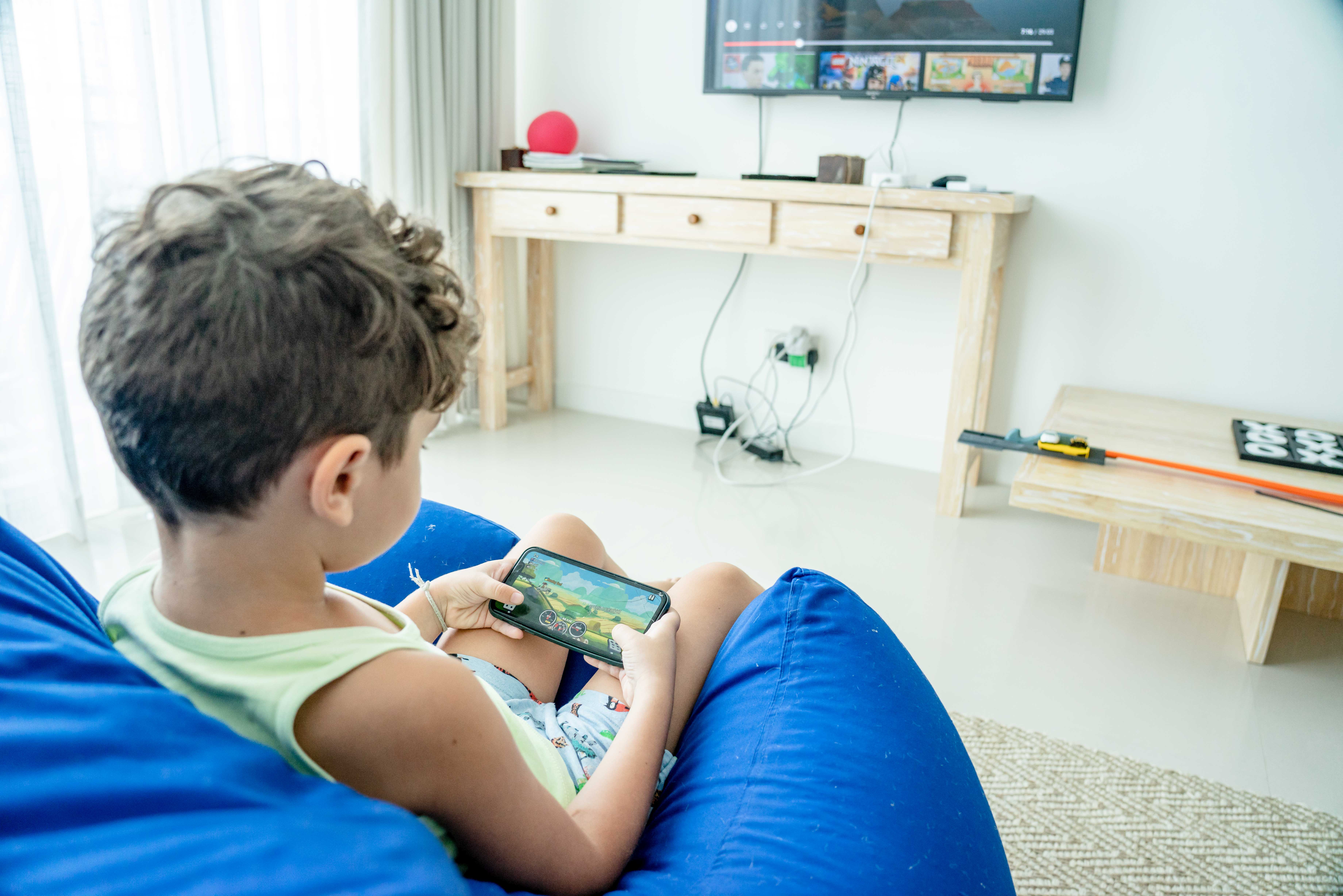 A kid playing a mobile game on a smartphone, with a TV displaying a gaming interface on the wall in the background.
