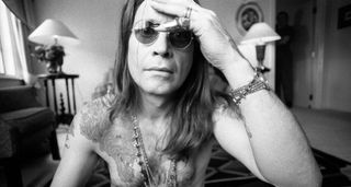 A black-and-white portrait of a shirtless Ozzy Osbourne in John Lennon sunglasses and his hand on his forehead.